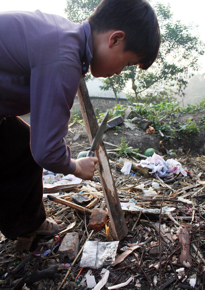 Ở ngoài sân, một em trong nhóm đang dùng dao đẽo một miếng củi làm thớt chặt thịt Ở ngoài sân, một em trong nhóm đang dùng dao đẽo một miếng củi làm thớt chặt thịt