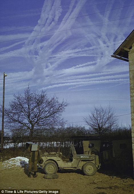 Máy bay của quân Đồng Minh để lại những vệt trắng xóa trên bầu trời, phía trên một chiếc xe một chiếc xe jeep đỗ bên ngoài một trang trại trong rừng Ardennes