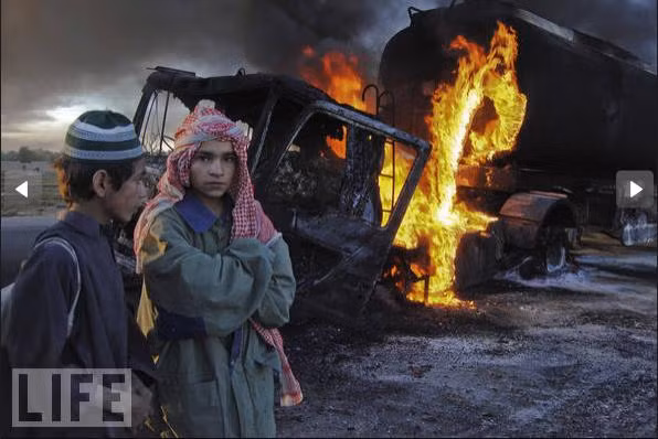 Hai học sinh đứng gần chiếc xe tải chờ nhiên liệu của NATO cháy đen sau cuộc tấn công của các tay súng tại tỉnh Baluchistan, Pakistan.