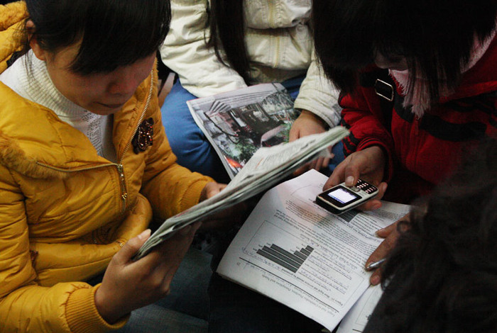 Một bạn gái đọc báo, bạn còn lại dùng điện thoại để tra từ điển và học tiếng anh.