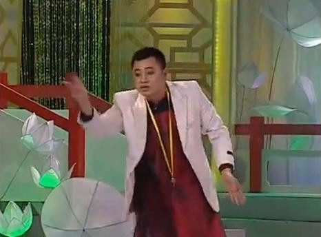 Tự Long trong trang phục áo the kết hợp áo vest, cổ đeo còi, tay vung đúng điệu bộ của bầu Kiên khi chỉ trích Liên đoàn bóng đá Việt Nam