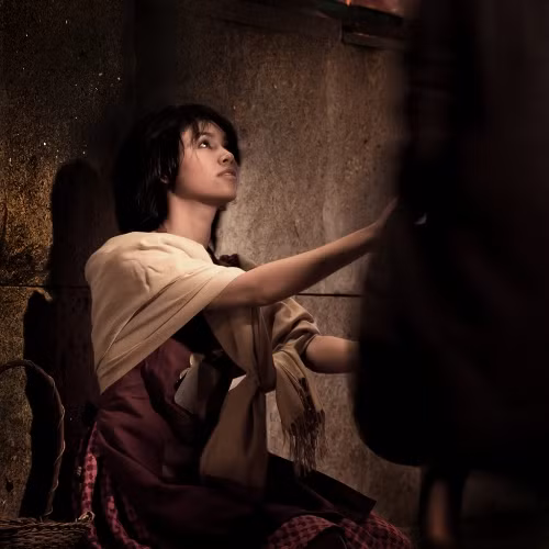 Rét dữ dội. Tuyết rơi. Trời đã tối hẳn. Đêm nay là đêm giao thừa. Giữa trời đông giá rét, một em gái nhỏ đầu trần, chân đi đất, đang dò dẫm trong đêm tối. Lúc ra khỏi nhà em có đi giày vải, nhưng giày vải phỏng có tác dụng gì kia chứ! Giày ấy của mẹ em để lại, rộng quá, em đã liên tiếp làm văng mất cả hai chiếc khi chạy qua đường, vào lúc hai chiếc xe ngựa đang phóng nước đại. Chiếc thứ nhất bị xe song mã nghiến, rồi dính theo tuyết vào bánh xe; thế là mất hút. Còn chiếc thứ hai, một thằng bé lượm được, cười sằng sặc, đem tung lên trời. Nó còn nói với em bé rằng nó sẽ giữ chiếc giày để làm nôi cho con chó sau này. Thế là em phải đi đất, chân em đỏ ửng lên, rồi tím bầm lại vì rét. Chiếc tạp dề cũ kỹ của em đựng đầy diêm và tay em còn cầm thêm một bao Em cố kiếm một nơi có nhiều người qua lại. Nhưng trời rét quá, khách qua đường đều rảo bước rất nhanh, chẳng ai đoái hoài đến lời chào hàng của em. Suốt ngày em chẳng bán được gì cả và chẳng ai bố thí cho em chút đỉnh. Em bé đáng thương vẫn lang thang trên đường. Cửa sổ mọi nhà đều sáng rực ánh đèn và trong phố sực nức mùi ngỗng quay. Chả là đêm giao thừa mà! Em tưởng nhớ lại năm xưa, khi bà nội hiền hậu của em còn sống, em cũng được đón giao thừa ở nhà. Nhưng thần chết đã đến cướp bà em đi mất, gia sản tiêu tán và gia đình em đã phải lìa ngôi nhà xinh xắn có dây trường xuân leo quanh, nơi em đã sống những ngày đầm ấm, để đến chui rúc trong một xó tối tăm, luôn luôn nghe những lời mắc nhiếc, chửi rủa... Em ngồi nép trong một góc tường, giữa hai ngôi nhà, một cái xây lùi lại một chút. Em thu đôi chân vào người, nhưng mỗi lúc em càng thấy rét buốt hơn. Tuy nhiên, em không thể nào về nhà nếu không bán được ít bao diêm, hay không ai bố thí cho một đồng xu nào đem về; nhất định là cha em sẽ đánh em. Vả lại, ở nhà cũng rét thế thôi. Cha con em ở trên gác, sát mái nhà và, mặc dầu đã nhét giẻ rách vào các kẽ hở trên vách, gió vẫn thổi rít vào trong nhà. Lúc này đôi bàn tay em đã cứng đờ ra. Chà! Giá quẹt một que diêm lên mà sưởi cho đỡ rét một chút nhỉ? Giá em có thể rút một que diêm ra quẹt vào tường mà hơ ngón tay nhỉ? Cuối cùng em đánh liều quẹt một que diêm. Diêm bén lửa thật là nhạy. Ngọn lửa lúc đầu xanh lam, dần dần biếc đi, trắng ra, rực hồng lên quanh que gỗ, sáng chói trông đến vui mắt. Em hơ đôi tay trên que diêm sáng rực như than hồng. Chà! Ánh sáng kỳ diệu làm sao! Em tưởng chừng như đang ngồi trước một lò sưởi bằng sắt có những hình nổi bằng đồng bóng nhoáng. Trong lò, lửa cháy nom đến vui mắt và tỏa ra hơi nóng dịu dàng. Em quẹt que diêm thứ hai, diêm cháy và sáng rực lên. Bức tường như biến thành một tấm rèm bằng vải màn. Em nhìn thấu tận trong nhà. Bàn ăn đã dọn, khăn trải trắng tinh... ... trên bàn bày toàn bát đĩa bằng sứ quý giá, và có cả một con ngỗng quay. Nhưng điều kỳ diệu nhất là ngỗng ta nhảy ra khỏi dĩa và mang cả dao ăn, phóng sết, cắm trên lưng, tiến về phía em bé. Em quẹt que diêm thứ ba. Bỗng em thấy hiện ra một cây thông Noel. Cây này lớn và trang trí lộng lẫy hơn cây mà em đã được thấy năm ngoái qua cửa kính một nhà buôn giàu có... Em với đôi tay về phía cây... nhưng diêm tắt.... Em quẹt một que diêm nữa vào tường, một ánh sáng xanh tỏa ra xung quanh và em bé nhìn thấy rõ ràng là bà em đang mỉm cười với em. - Bà ơi! Em bé reo lên, cho cháu đi với! Cháu biết rằng diêm tắt thì bà cũng biến đi mất như lò sưởi, ngỗng quay và cây Noel ban nãy, nhưng xin bà đừng bỏ cháu ở nơi này; trước kia, khi bà chưa về với thượng đế chí nhân, bà cháu ta đã từng sung sướng biết bao! Dạo ấy bà đã từng nhủ cháu rằng nếu cháu ngoan ngoãn cháu sẽ được gặp lại bà: Bà ơi, cháu van bà, bà xin với Thượng Đế chí nhân cho cháu về với bà. Chắc Người không từ chối đâu. Que diêm tắt phụt và ảo ảnh rực sáng trên khuôn mặt em bé cũng biến mất. Thế là em quẹt tất cả những que diêm còn lại trong bao. Em muốn níu bà em lại! Diêm nối nhau chiếu sáng như giữa ban ngày. Chưa bao giờ em thấy bà em to lớn và đẹp lão như thế này. Bà cụ cầm lấy tay em, rồi hai bà cháu bay vụt lên cao, cao mãi, chẳng còn đói rét, đau buồn nào đe dọa họ nữa. Họ đã về với Thuợng đế. Sáng hôm sau, tuyết vẫn phủ kín mặt đất, nhưng mặt trời lên, trong sáng, chói chang trên bầu trời xanh nhợt. Mọi người vui vẻ ra khỏi nhà...Trong buổi sáng lạnh lẽo ấy, ở một xóa tường người ta thấy một em gái có đôi má hồng... ... và đôi môi đang mỉm cười. Em đã chết vì giá rét trong đêm giao thừa. Rét dữ dội. Tuyết rơi. Trời đã tối hẳn. Đêm nay là đêm giao thừa. Giữa trời đông giá rét, một em gái nhỏ đầu trần, chân đi đất, đang dò dẫm trong đêm tối. Lúc ra khỏi nhà em có đi giày vải, nhưng giày vải phỏng có tác dụng gì kia chứ! Giày ấy của mẹ em để lại, rộng quá, em đã liên tiếp làm văng mất cả hai chiếc khi chạy qua đường, vào lúc hai chiếc xe ngựa đang phóng nước đại. Chiếc thứ nhất bị xe song mã nghiến, rồi dính theo tuyết vào bánh xe; thế là mất hút. Còn chiếc thứ hai, một thằng bé lượm được, cười sằng sặc, đem tung lên trời. Nó còn nói với em bé rằng nó sẽ giữ chiếc giày để làm nôi cho con chó sau này. Thế là em phải đi đất, chân em đỏ ửng lên, rồi tím bầm lại vì rét. Chiếc tạp dề cũ kỹ của em đựng đầy diêm và tay em còn cầm thêm một bao Em cố kiếm một nơi có nhiều người qua lại. Nhưng trời rét quá, khách qua đường đều rảo bước rất nhanh, chẳng ai đoái hoài đến lời chào hàng của em. Suốt ngày em chẳng bán được gì cả và chẳng ai bố thí cho em chút đỉnh. Em bé đáng thương vẫn lang thang trên đường. Cửa sổ mọi nhà đều sáng rực ánh đèn và trong phố sực nức mùi ngỗng quay. Chả là đêm giao thừa mà! Em tưởng nhớ lại năm xưa, khi bà nội hiền hậu của em còn sống, em cũng được đón giao thừa ở nhà. Nhưng thần chết đã đến cướp bà em đi mất, gia sản tiêu tán và gia đình em đã phải lìa ngôi nhà xinh xắn có dây trường xuân leo quanh, nơi em đã sống những ngày đầm ấm, để đến chui rúc trong một xó tối tăm, luôn luôn nghe những lời mắc nhiếc, chửi rủa... Em ngồi nép trong một góc tường, giữa hai ngôi nhà, một cái xây lùi lại một chút. Em thu đôi chân vào người, nhưng mỗi lúc em càng thấy rét buốt hơn. Tuy nhiên, em không thể nào về nhà nếu không bán được ít bao diêm, hay không ai bố thí cho một đồng xu nào đem về; nhất định là cha em sẽ đánh em. Vả lại, ở nhà cũng rét thế thôi. Cha con em ở trên gác, sát mái nhà và, mặc dầu đã nhét giẻ rách vào các kẽ hở trên vách, gió vẫn thổi rít vào trong nhà. Lúc này đôi bàn tay em đã cứng đờ ra. Chà! Giá quẹt một que diêm lên mà sưởi cho đỡ rét một chút nhỉ? Giá em có thể rút một que diêm ra quẹt vào tường mà hơ ngón tay nhỉ? Cuối cùng em đánh liều quẹt một que diêm. Diêm bén lửa thật là nhạy. Ngọn lửa lúc đầu xanh lam, dần dần biếc đi, trắng ra, rực hồng lên quanh que gỗ, sáng chói trông đến vui mắt. Em hơ đôi tay trên que diêm sáng rực như than hồng. Chà! Ánh sáng kỳ diệu làm sao! Em tưởng chừng như đang ngồi trước một lò sưởi bằng sắt có những hình nổi bằng đồng bóng nhoáng. Trong lò, lửa cháy nom đến vui mắt và tỏa ra hơi nóng dịu dàng. Em quẹt que diêm thứ hai, diêm cháy và sáng rực lên. Bức tường như biến thành một tấm rèm bằng vải màn. Em nhìn thấu tận trong nhà. Bàn ăn đã dọn, khăn trải trắng tinh... ... trên bàn bày toàn bát đĩa bằng sứ quý giá, và có cả một con ngỗng quay. Nhưng điều kỳ diệu nhất là ngỗng ta nhảy ra khỏi dĩa và mang cả dao ăn, phóng sết, cắm trên lưng, tiến về phía em bé. Em quẹt que diêm thứ ba. Bỗng em thấy hiện ra một cây thông Noel. Cây này lớn và trang trí lộng lẫy hơn cây mà em đã được thấy năm ngoái qua cửa kính một nhà buôn giàu có... Em với đôi tay về phía cây... nhưng diêm tắt.... Em quẹt một que diêm nữa vào tường, một ánh sáng xanh tỏa ra xung quanh và em bé nhìn thấy rõ ràng là bà em đang mỉm cười với em. - Bà ơi! Em bé reo lên, cho cháu đi với! Cháu biết rằng diêm tắt thì bà cũng biến đi mất như lò sưởi, ngỗng quay và cây Noel ban nãy, nhưng xin bà đừng bỏ cháu ở nơi này; trước kia, khi bà chưa về với thượng đế chí nhân, bà cháu ta đã từng sung sướng biết bao! Dạo ấy bà đã từng nhủ cháu rằng nếu cháu ngoan ngoãn cháu sẽ được gặp lại bà: Bà ơi, cháu van bà, bà xin với Thượng Đế chí nhân cho cháu về với bà. Chắc Người không từ chối đâu. Que diêm tắt phụt và ảo ảnh rực sáng trên khuôn mặt em bé cũng biến mất. Thế là em quẹt tất cả những que diêm còn lại trong bao. Em muốn níu bà em lại! Diêm nối nhau chiếu sáng như giữa ban ngày. Chưa bao giờ em thấy bà em to lớn và đẹp lão như thế này. Bà cụ cầm lấy tay em, rồi hai bà cháu bay vụt lên cao, cao mãi, chẳng còn đói rét, đau buồn nào đe dọa họ nữa. Họ đã về với Thuợng đế. Sáng hôm sau, tuyết vẫn phủ kín mặt đất, nhưng mặt trời lên, trong sáng, chói chang trên bầu trời xanh nhợt. Mọi người vui vẻ ra khỏi nhà...Trong buổi sáng lạnh lẽo ấy, ở một xóa tường người ta thấy một em gái có đôi má hồng... ... và đôi môi đang mỉm cười. Em đã chết vì giá rét trong đêm giao thừa.