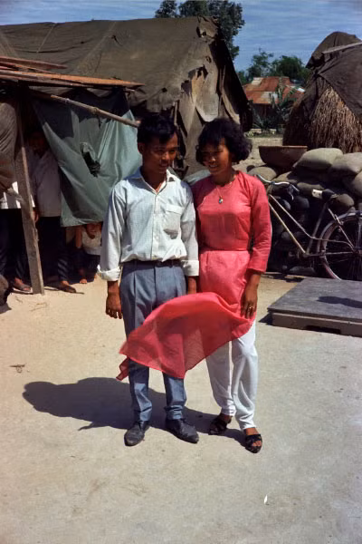 Mọi người rót cho tôi một chất lỏng từ chai Pepsi cũ. Nó khiến tôi cháy cổ họng sau khi uống một ngụm. Điệu bộ của tôi có lẽ giống như một thằng hề, và người Việt Nam đổ lại thứ chất lỏng gắt như axit này vào chai. Sau đó họ pha chúng với Pepsi và lại mời tôi uống. Nhưng phản ứng của tôi cũng không khá hơn lần trước. Ảnh: Cô dâu và chú rể ngượng nghịu chụp ảnh kỷ niệm. Sau lưng họ là những bao cát quân sự.