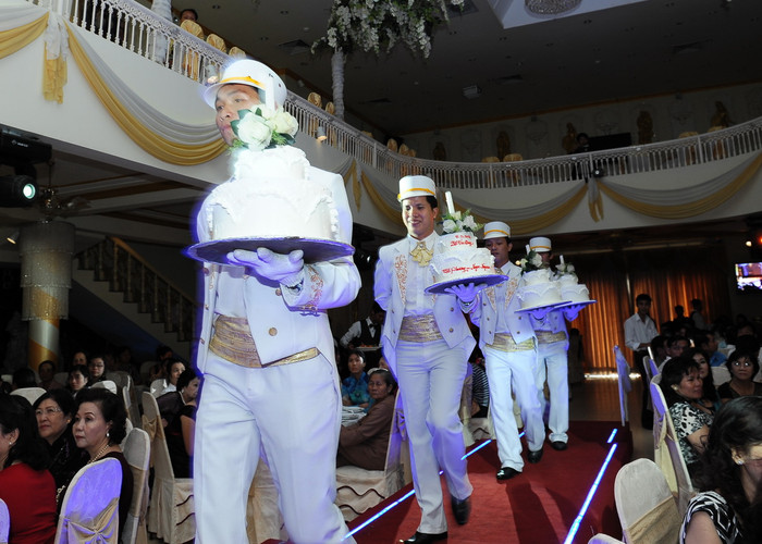 5 chiếc bánh kem được mang ra sân khấu bởi 5 nhân viên lễ tân cao lớn.