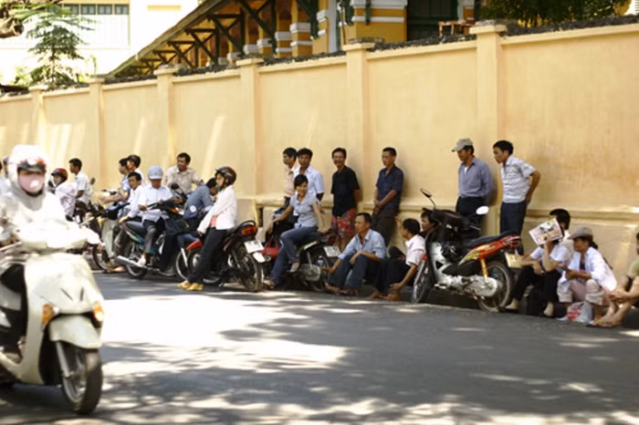 Phụ huynh ngồi xếp hàng cạnh tường rào nhà trường chờ con làm bài thi.